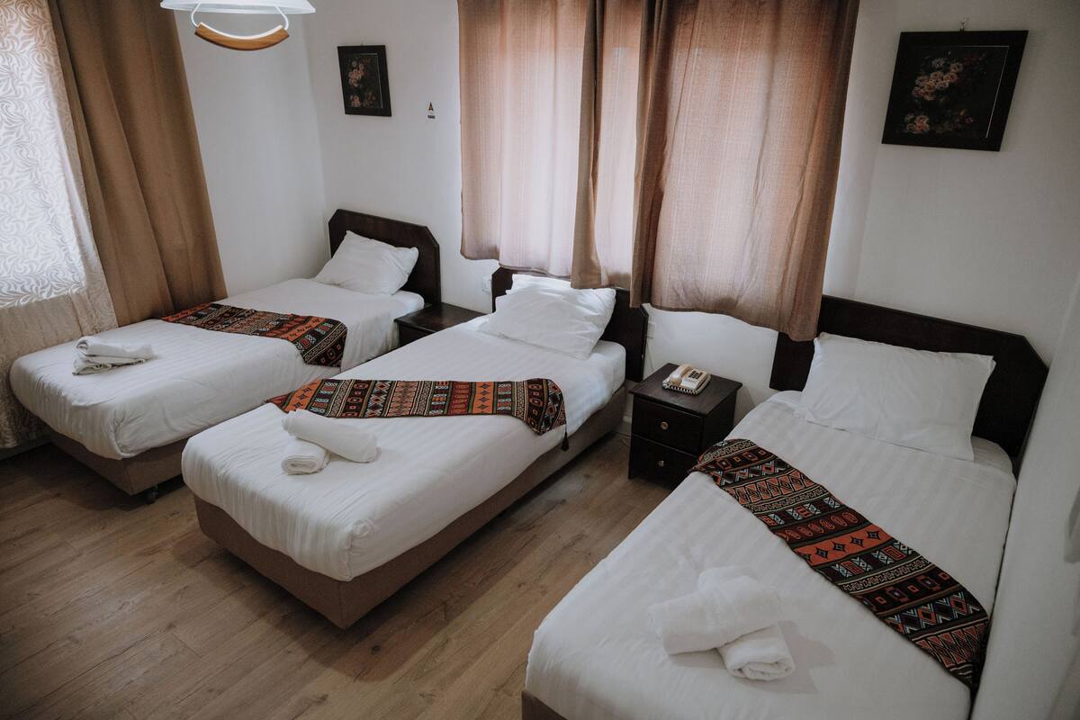 A hotel room with three single beds, wooden flooring, and a nightstand with a phone.