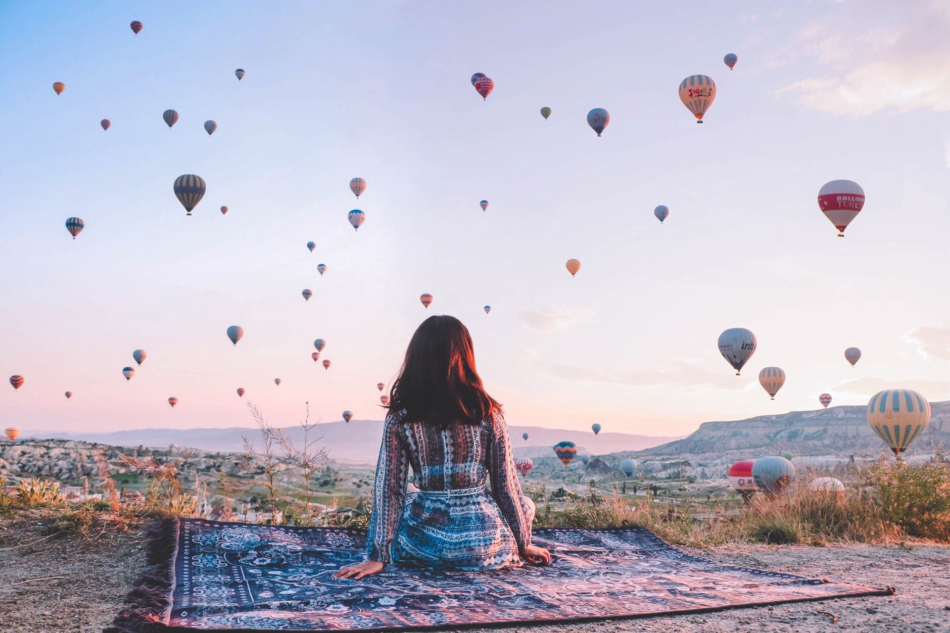 A person sitting on a patterned rug outdoors with a view of a landscape and hot air balloons in the sky.