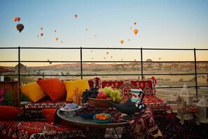Un balcon avec un canapé, des coussins, une table garnie de fruits et une théière, et une vue sur des montgolfières.