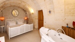 A bedroom with a white bed, a large arched stone ceiling, a wooden door, and a bathtub.