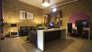 Een moderne woonkamer met een stenen muur, een zwarte leren bank, een witte salontafel, een televisie en een bureau met stoelen.