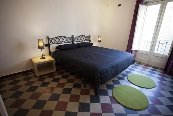A bedroom with a checkered floor, a bed with a dark headboard, two bedside lamps, and a window with curtains.