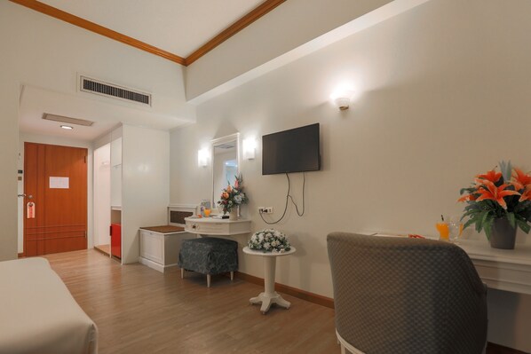 A hotel room with a wooden floor, a bed, a desk with a chair, a small table with flowers, a TV mounted on the wall, and a door leading to another room.