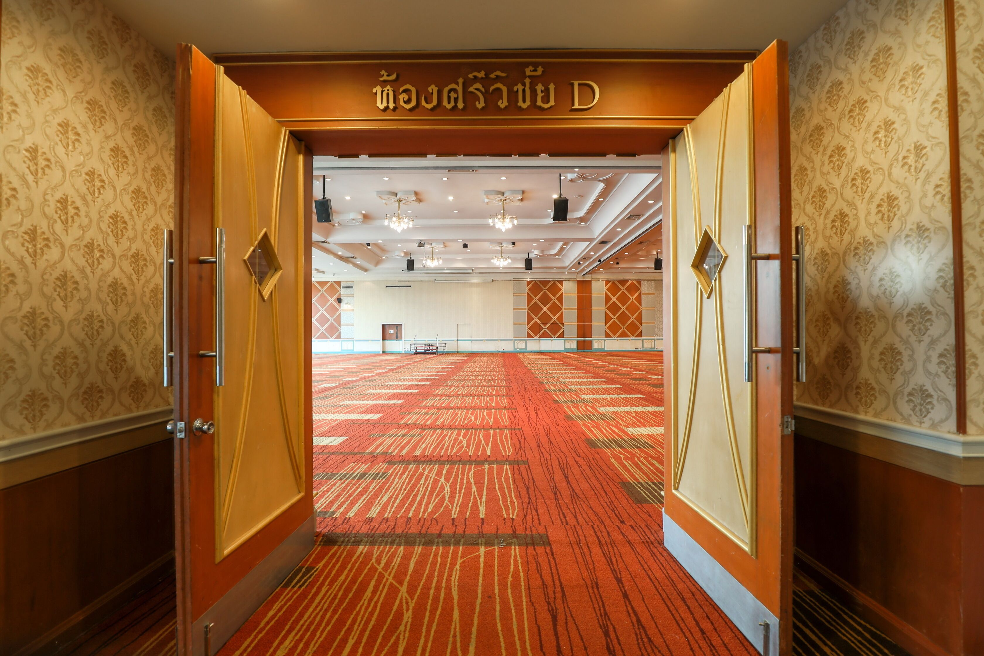 Vista del interior de un vestíbulo de hotel con papel tapiz estampado, alfombra roja y un letrero con escritura tailandesa.