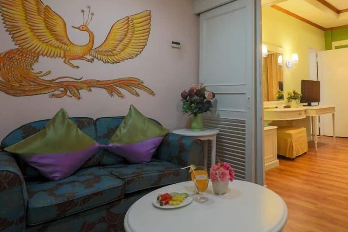 A living room with a sofa, a round coffee table with fruits and drinks, a painting of a bird, and a view into a dining area with a TV and chairs.