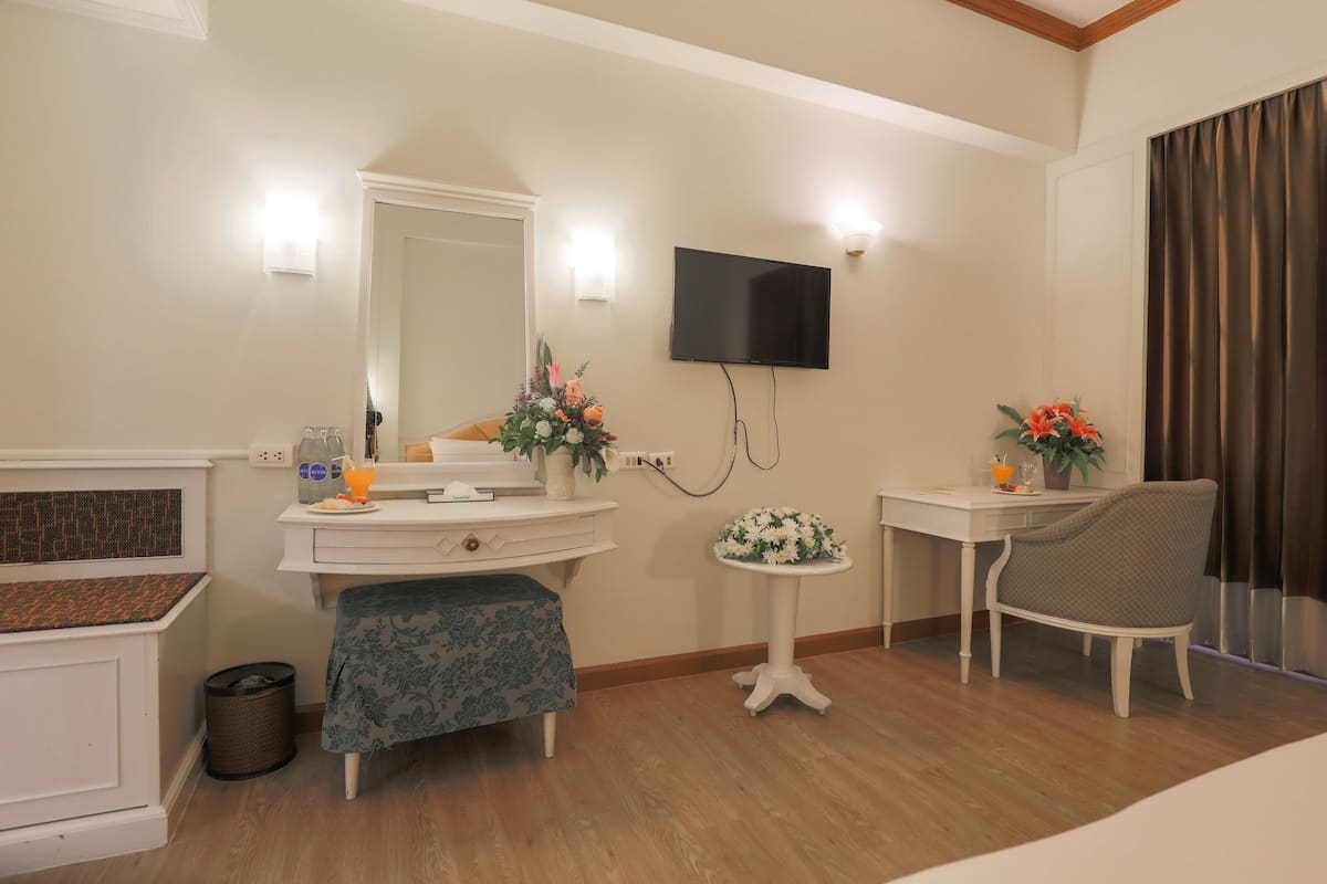 A hotel room with a wooden floor, a white vanity with a mirror, a flat-screen TV mounted on the wall, a small round table with a flower vase, and a chair.