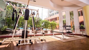 A gym with exercise equipment, large windows, and a view of a building and trees. - Wangtai Hotel (Surat Thani)