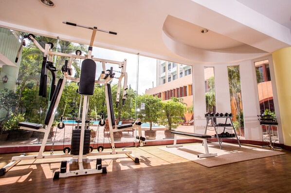 A gym with exercise equipment, large windows, and a view of a building and trees. - Wangtai Hotel (Surat Thani)