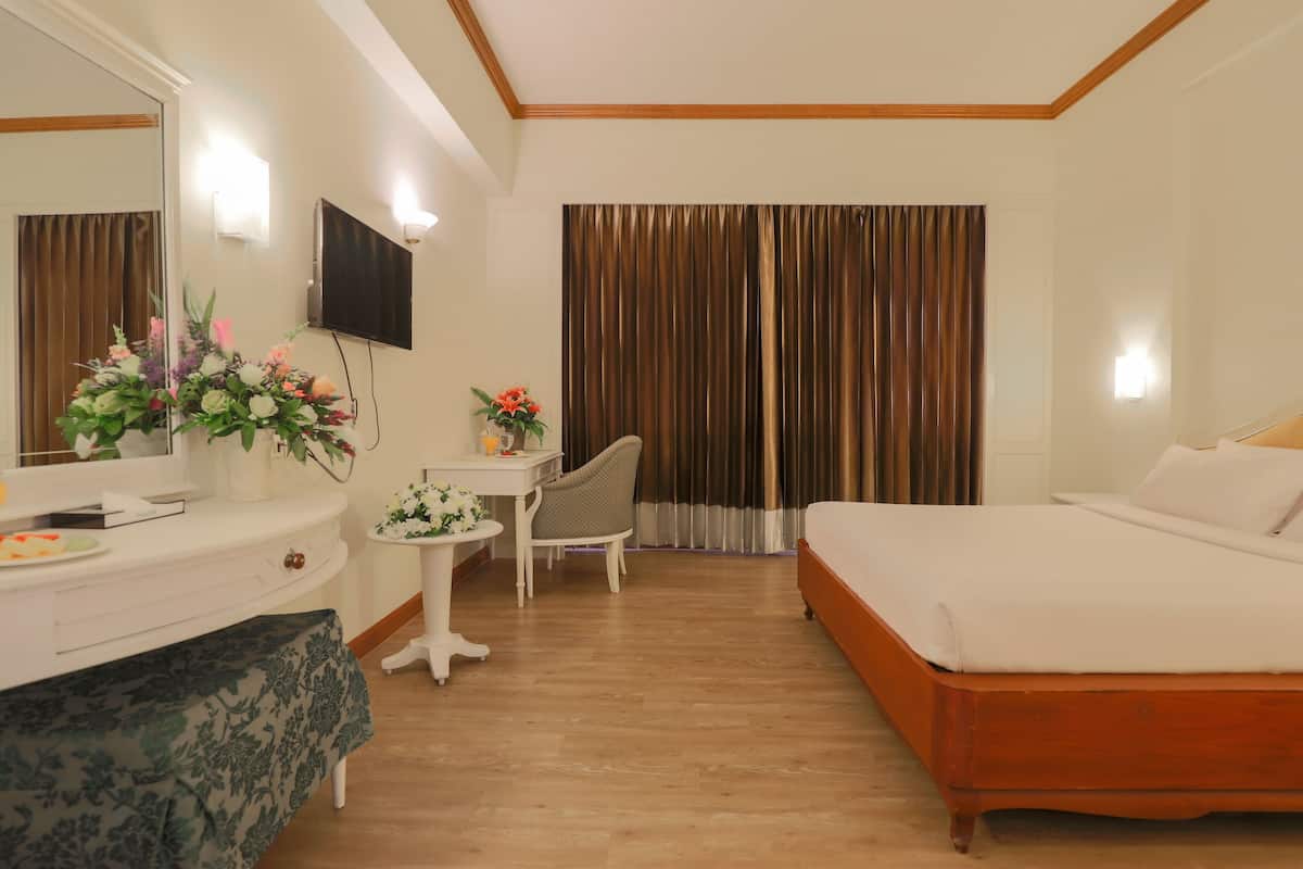 A hotel room with a bed, a desk with a chair, a small table with a vase of flowers, a mirror, and a TV on the wall.