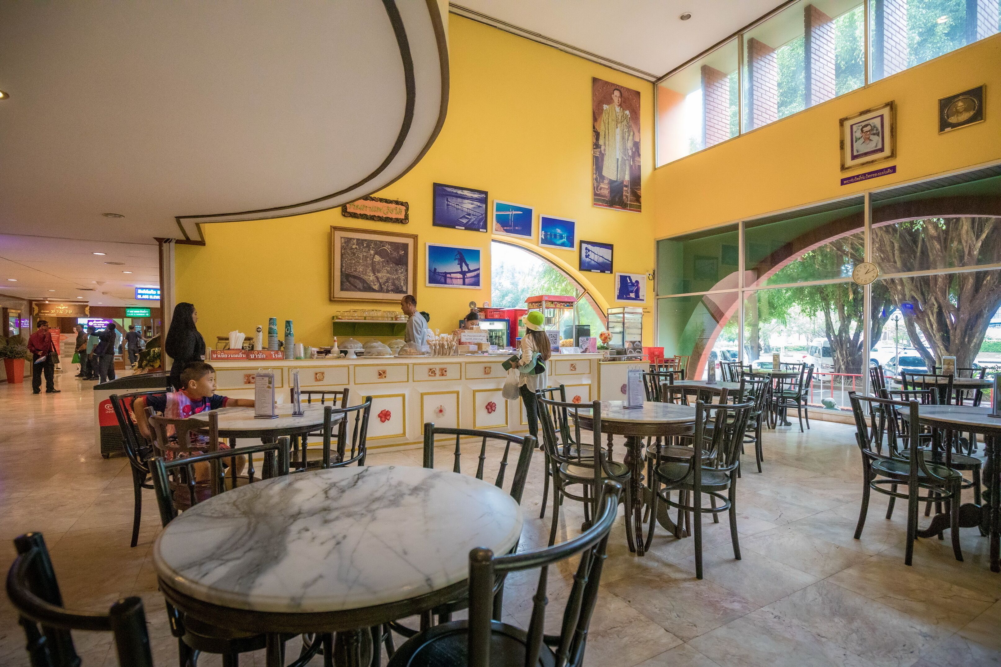 A spacious indoor dining area with marble tables and wooden chairs, featuring a yellow wall with framed pictures and a large window with a view of trees.