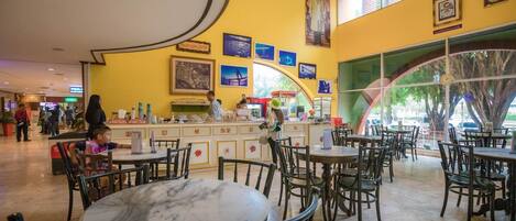 A spacious indoor dining area with marble tables and wooden chairs, featuring a yellow wall with framed pictures and a large window with a view of trees.