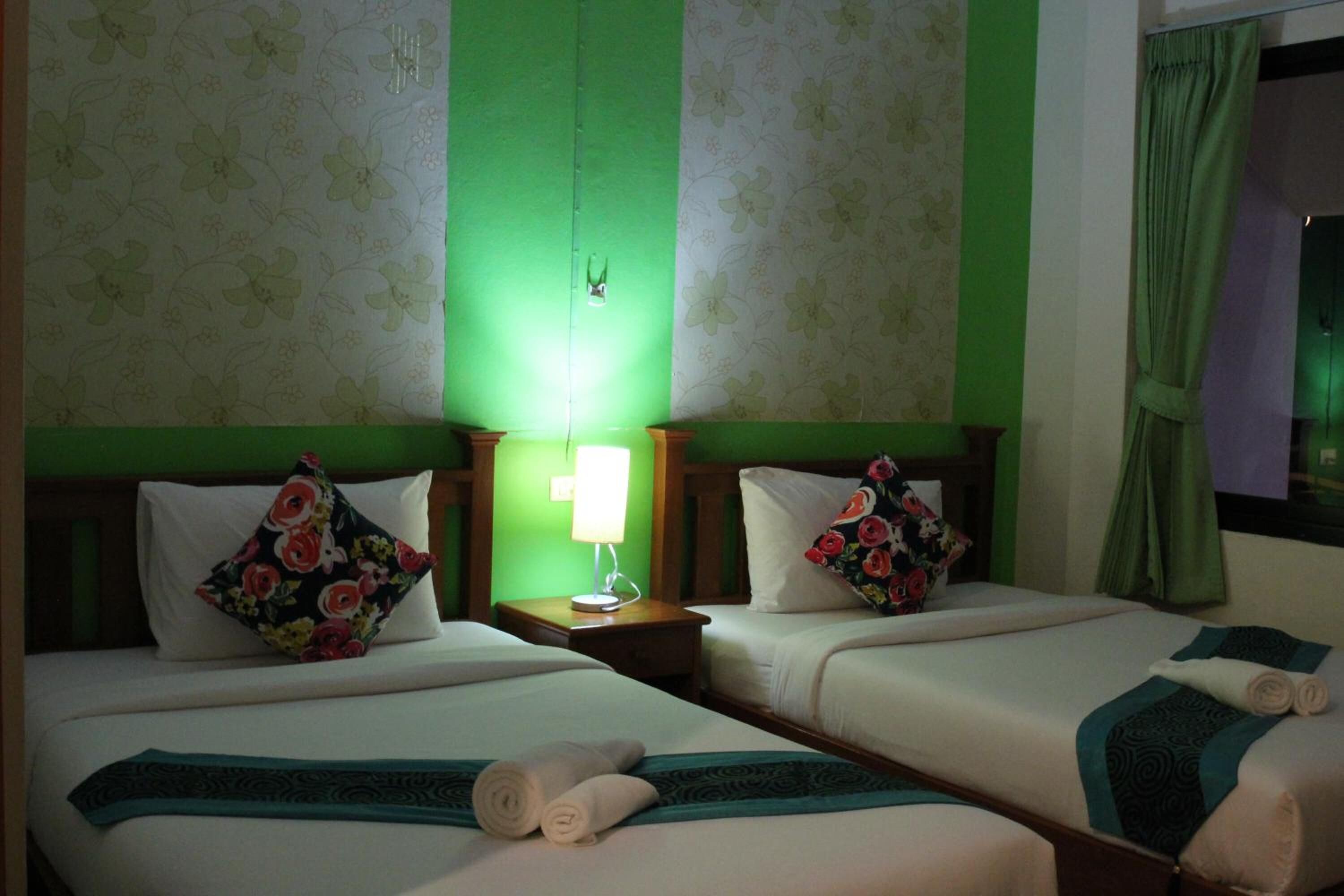 Habitación de hotel con dos camas, cada una con un cojín floral y una colcha estampada en tonos aguamarina. Hay una mesita de noche con una lámpara entre las camas.