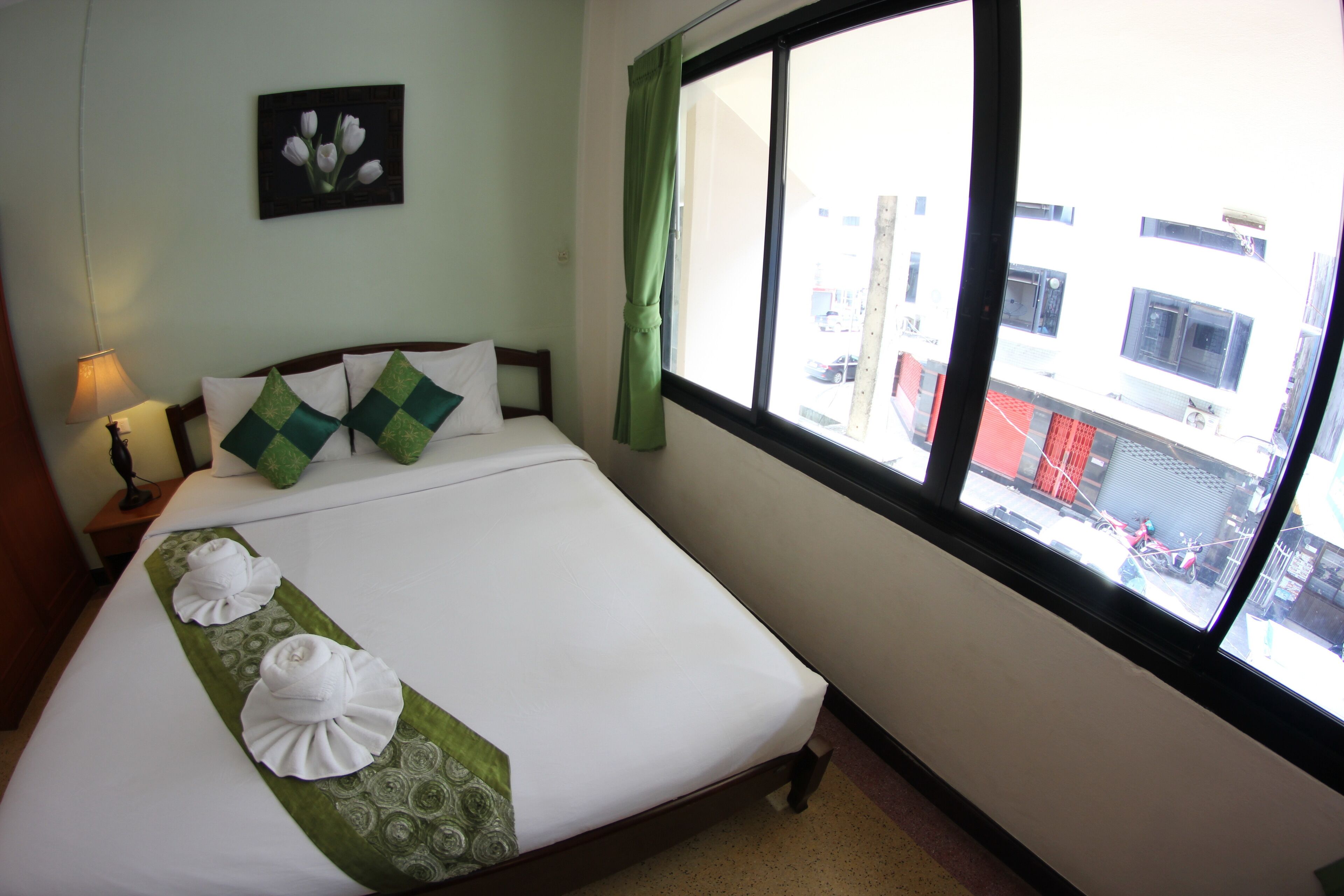 Una cama bien tendida con un corredor verde y almohadas blancas, una lámpara de noche y una ventana con vistas a edificios y un vehículo rojo fuera.