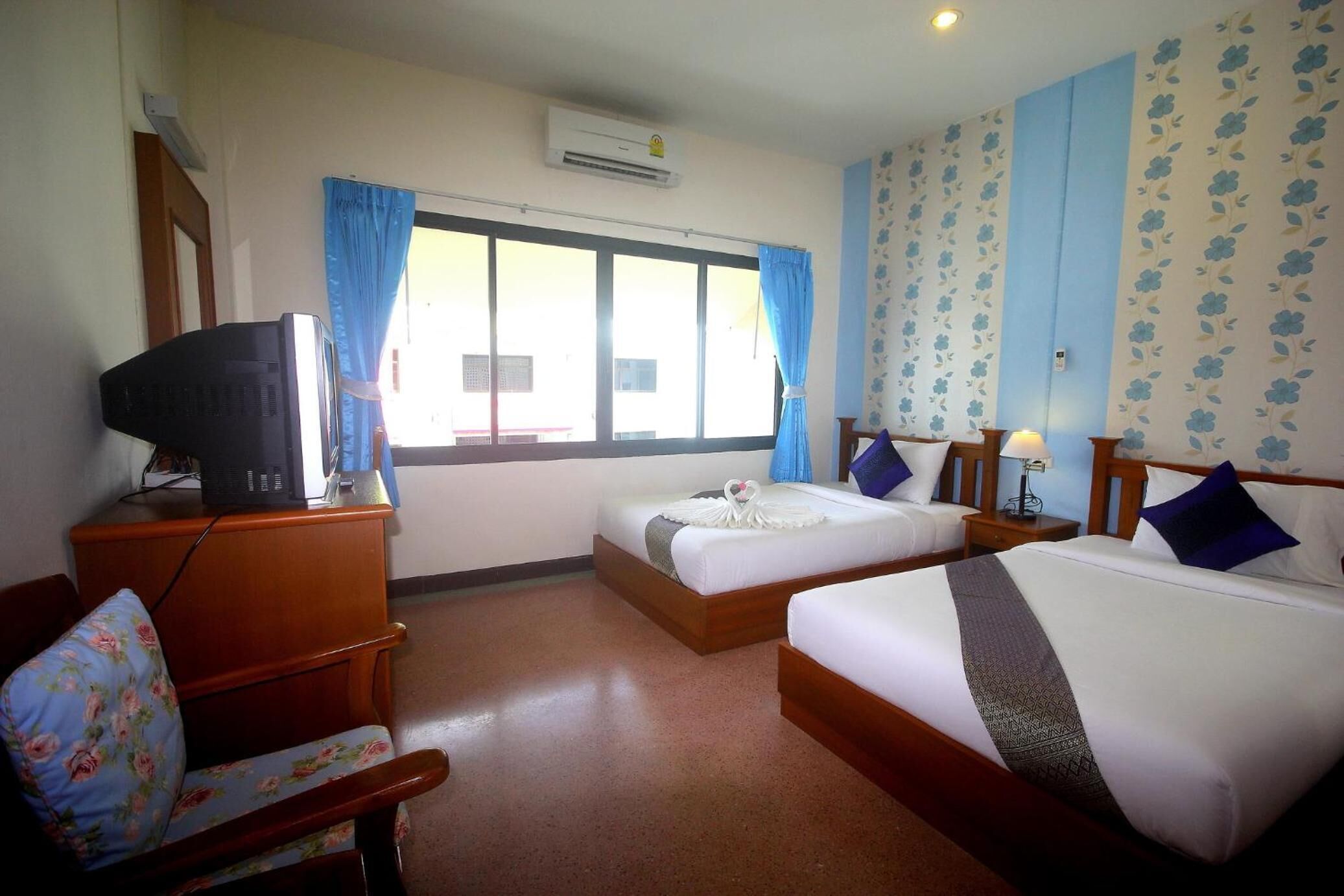 A hotel room with two beds, a television on a wooden dresser, a chair with floral upholstery, and a window with blue curtains.