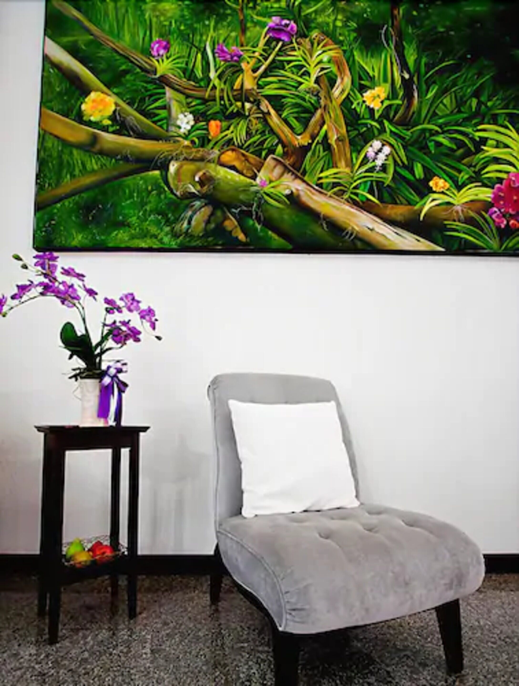 A room with a grey armchair, a small table with a potted plant, and a large floral painting on the wall.