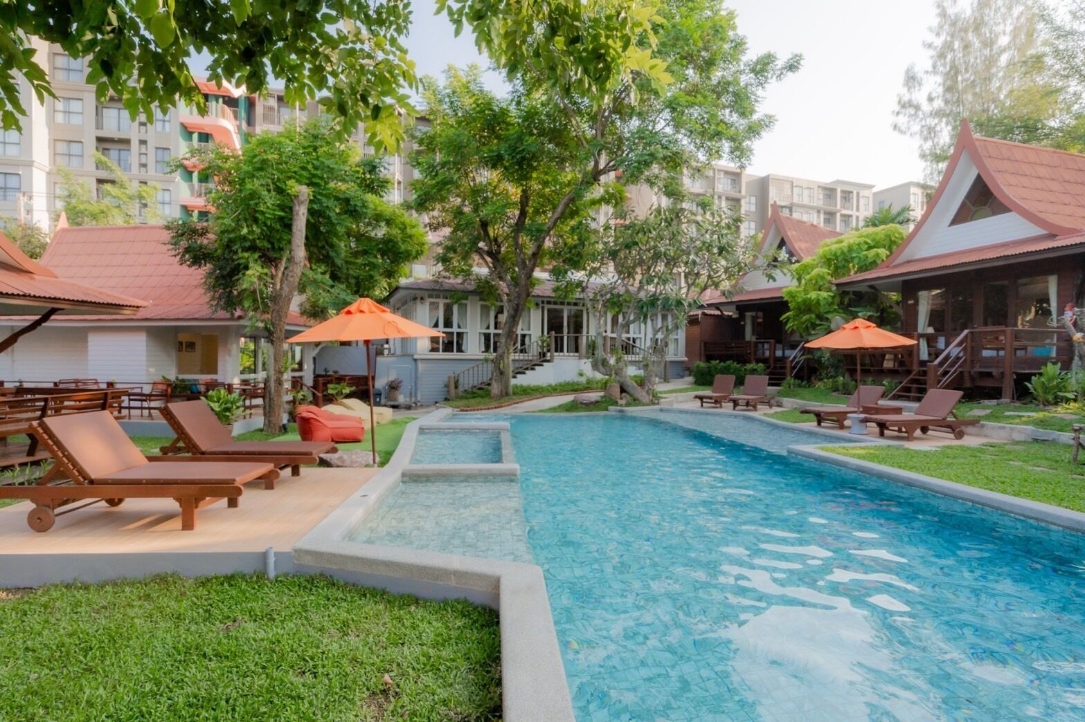 Una piscina con sillas de descanso y sombrillas, rodeada de vegetación y edificios.