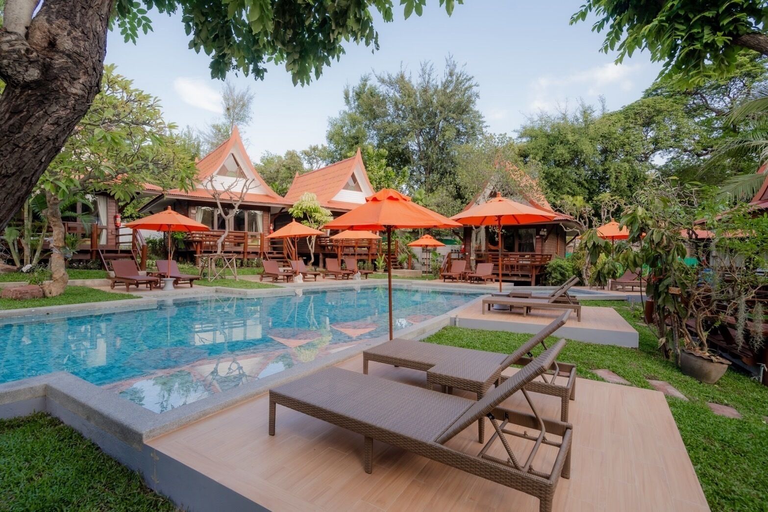A poolside area with lounge chairs and umbrellas, surrounded by greenery and buildings with red roofs.
