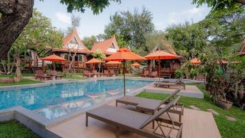 A poolside area with lounge chairs and umbrellas, surrounded by greenery and buildings with red roofs.