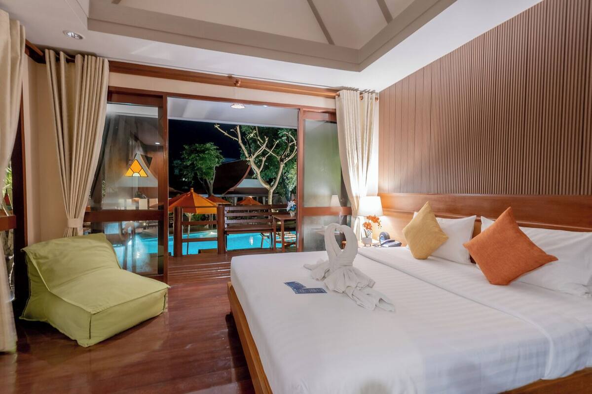 A hotel room with a bed, a green chair, a pool view, and a wooden ceiling.