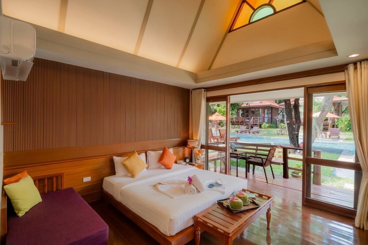 A hotel room with a large bed, a wooden headboard, a small table with a tray of fruit, and a view of an outdoor pool area.