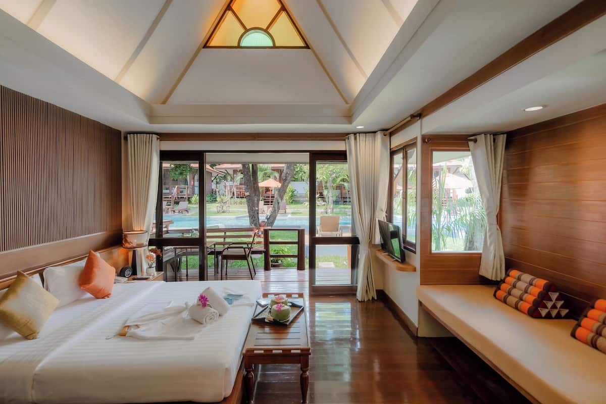 A hotel room with a bed, a TV, a sofa, and a view of a pool and garden.