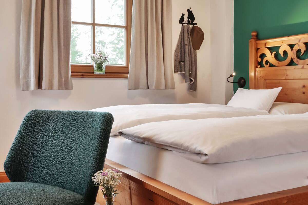 Un dormitorio con una cama de madera, una silla verde, una mesita con un jarrón y una ventana con cortinas.