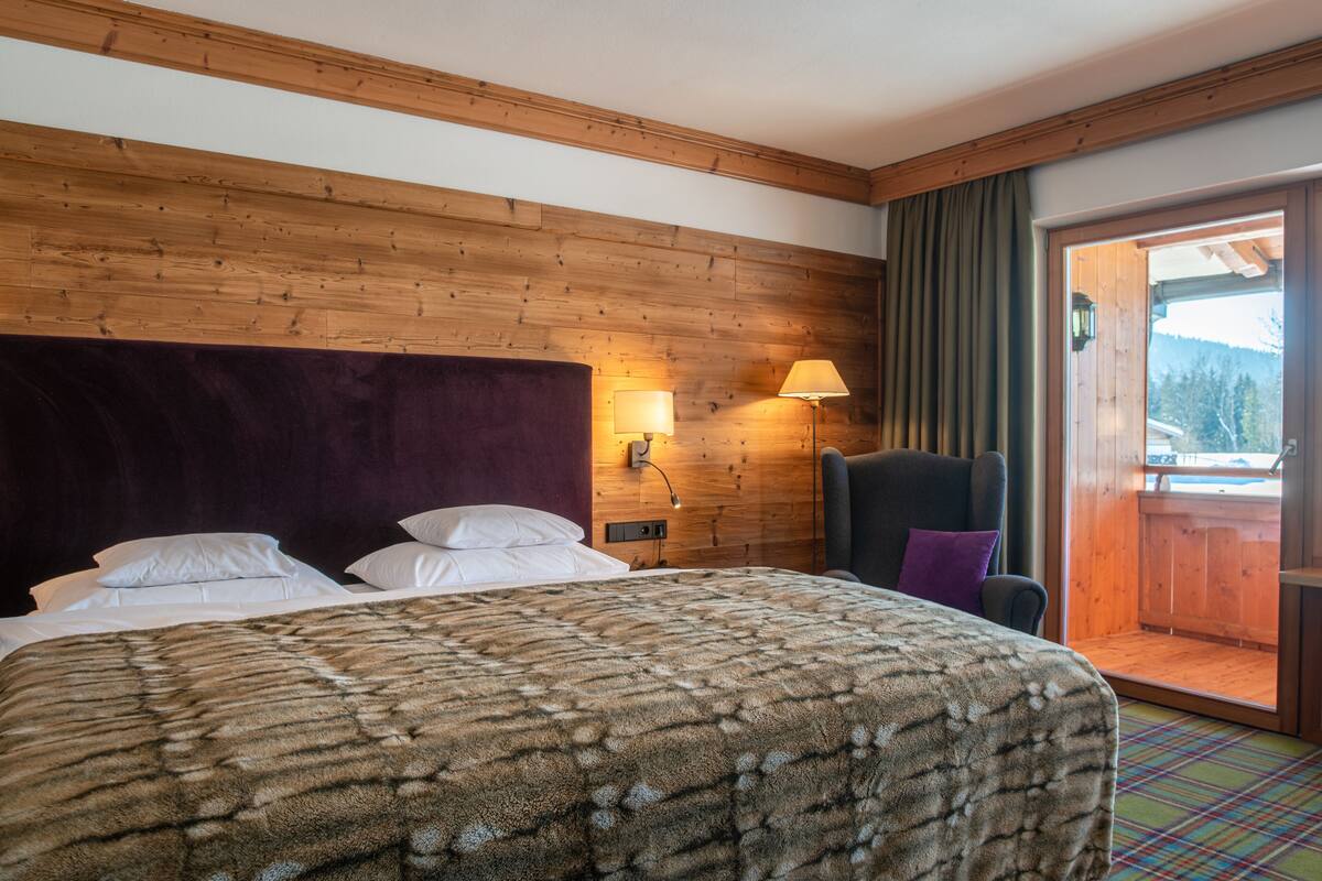 Habitación de hotel con cabecera de madera, cama grande con manta estampada, silla con cojín morado, lámpara de pared y vista a una terraza de madera a través de una puerta de cristal.