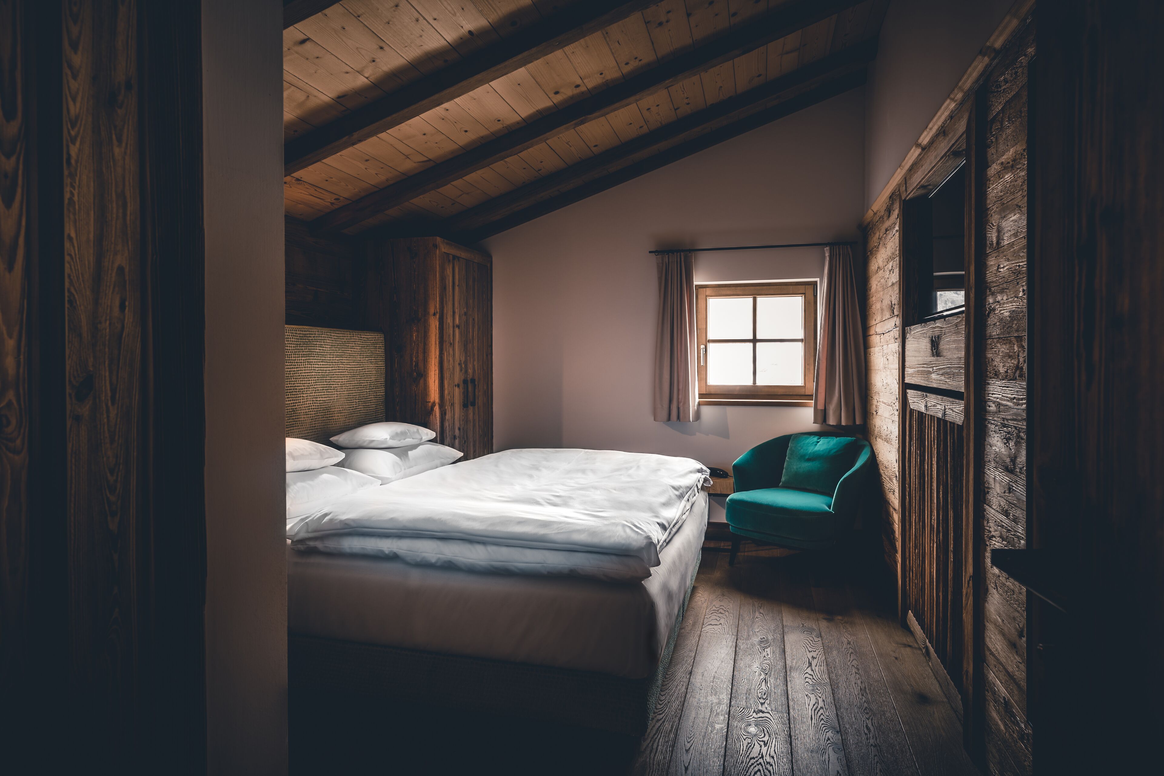 A cozy bedroom with a wooden ceiling, a bed with white linens, a green chair, and a window with curtains.