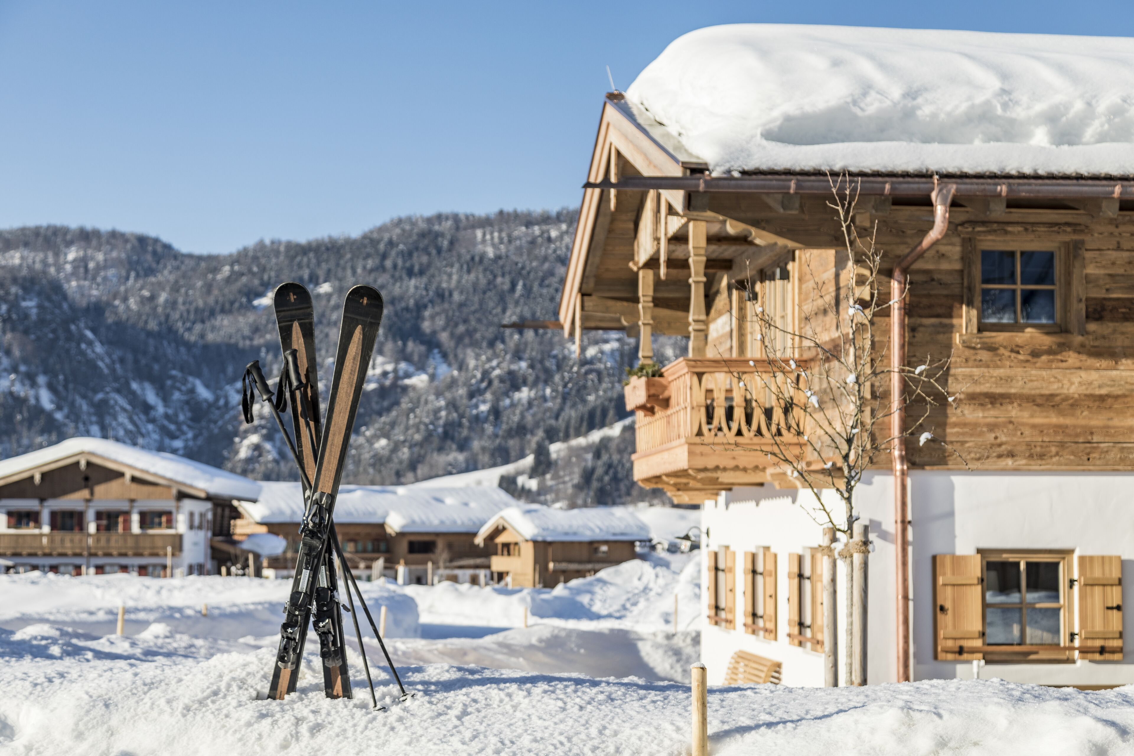 A wooden chalet with a balcony and snow-covered roof, surrounded by other similar buildings and a mountainous backdrop.