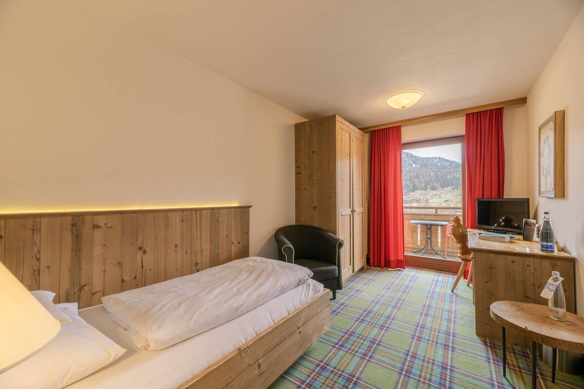 Habitación de hotel con cama de madera, un escritorio con computadora, una silla y vistas a la montaña a través de una ventana con cortinas rojas.