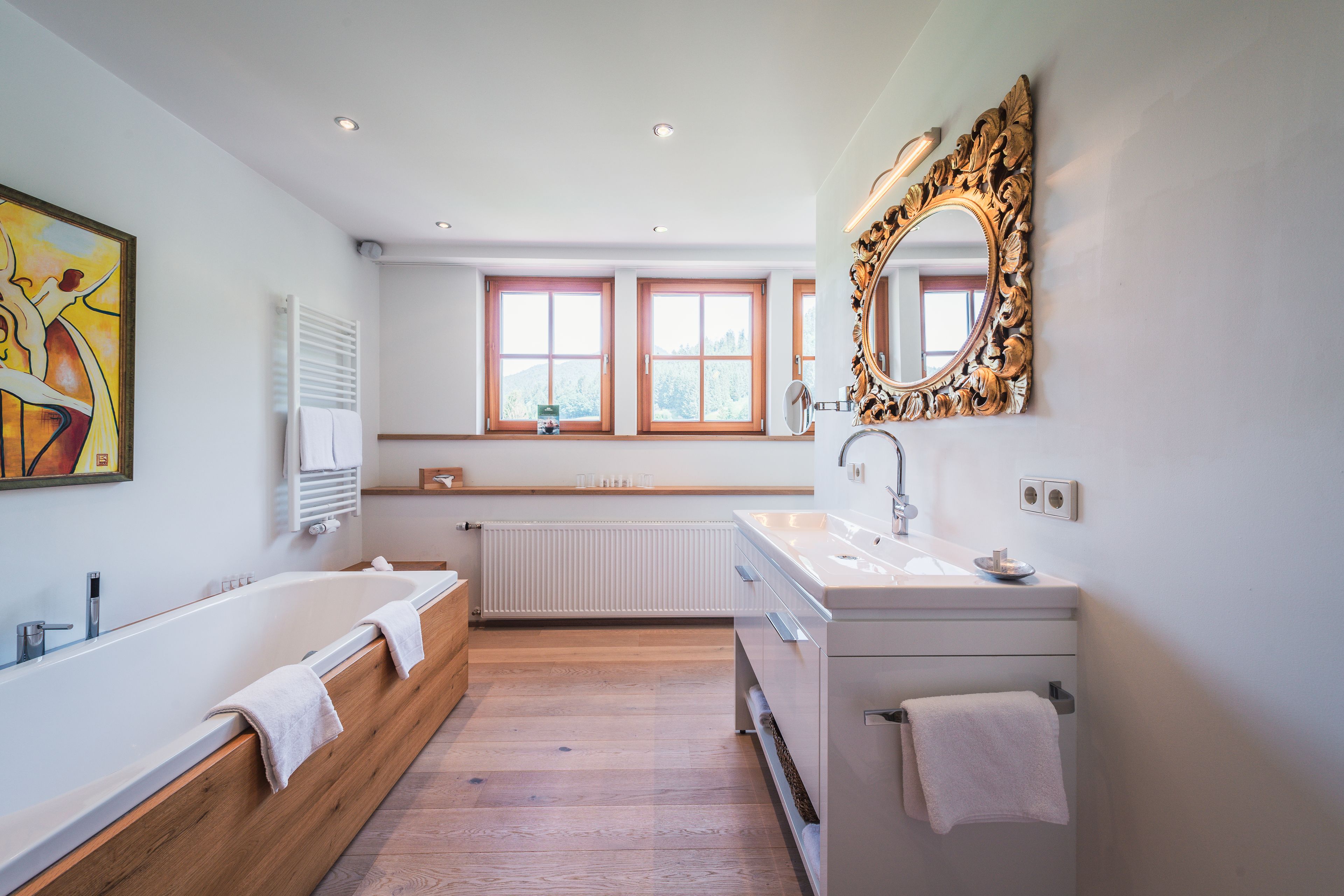 A modern bathroom with a bathtub, a sink, and a large mirror. There are windows with wooden frames and a radiator.