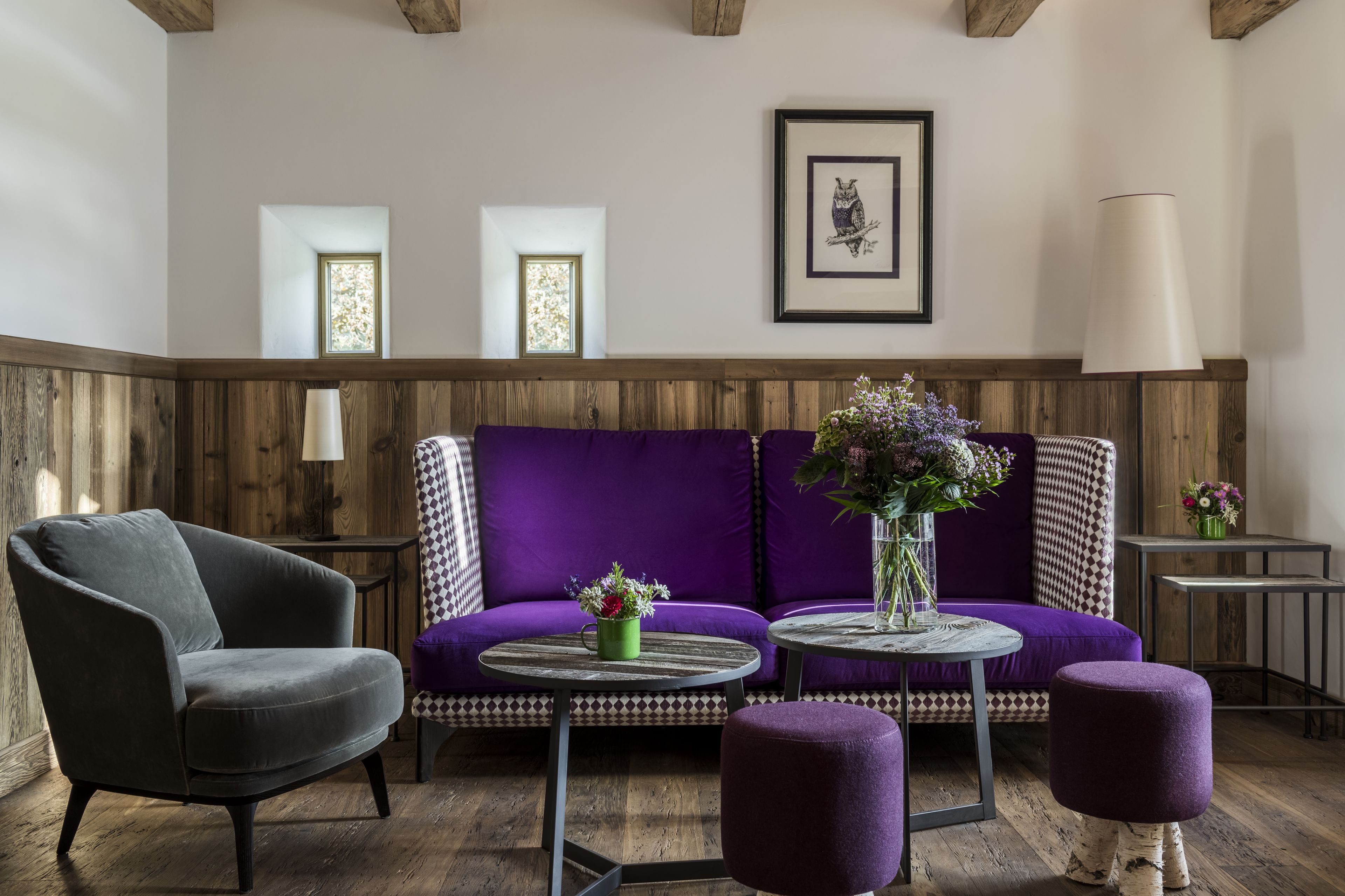 Ein Zimmer mit einem violetten Sofa, einem grauen Sessel und einem runden Tisch mit Blumen in einer Vase. Es gibt zwei Fenster mit verzierten Rahmen und ein gerahmtes Bild an der Wand.