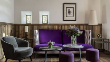 Ein Zimmer mit einem violetten Sofa, einem grauen Sessel und einem runden Tisch mit Blumen in einer Vase. Es gibt zwei Fenster mit verzierten Rahmen und ein gerahmtes Bild an der Wand.