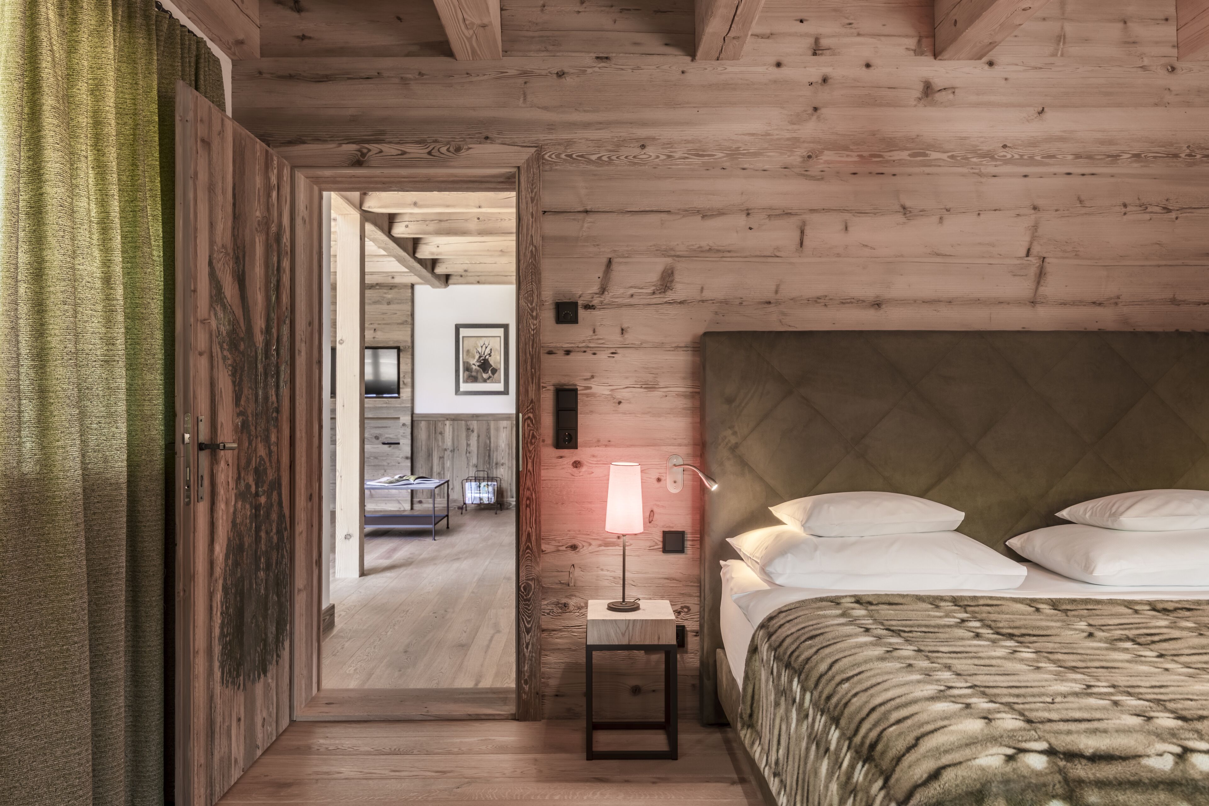 A wooden bedroom with a bed, bedside lamp, and a view into another room.