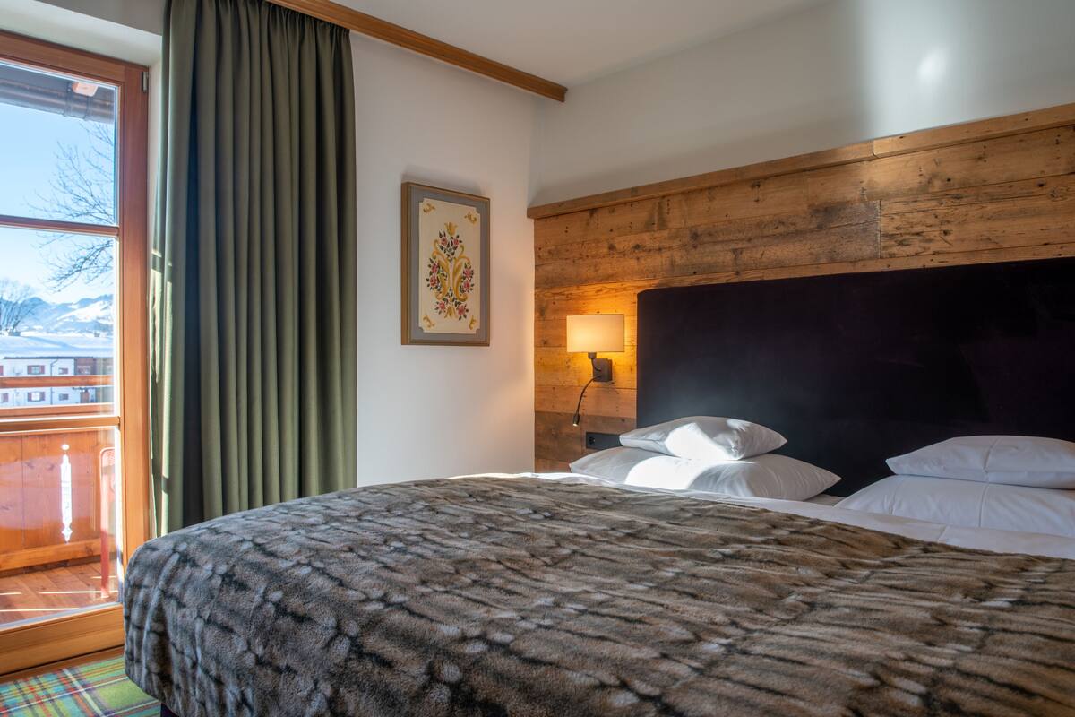 Habitación de hotel con cama, ventana con cortinas, cabecero de madera, lámpara de pared e imagen enmarcada.