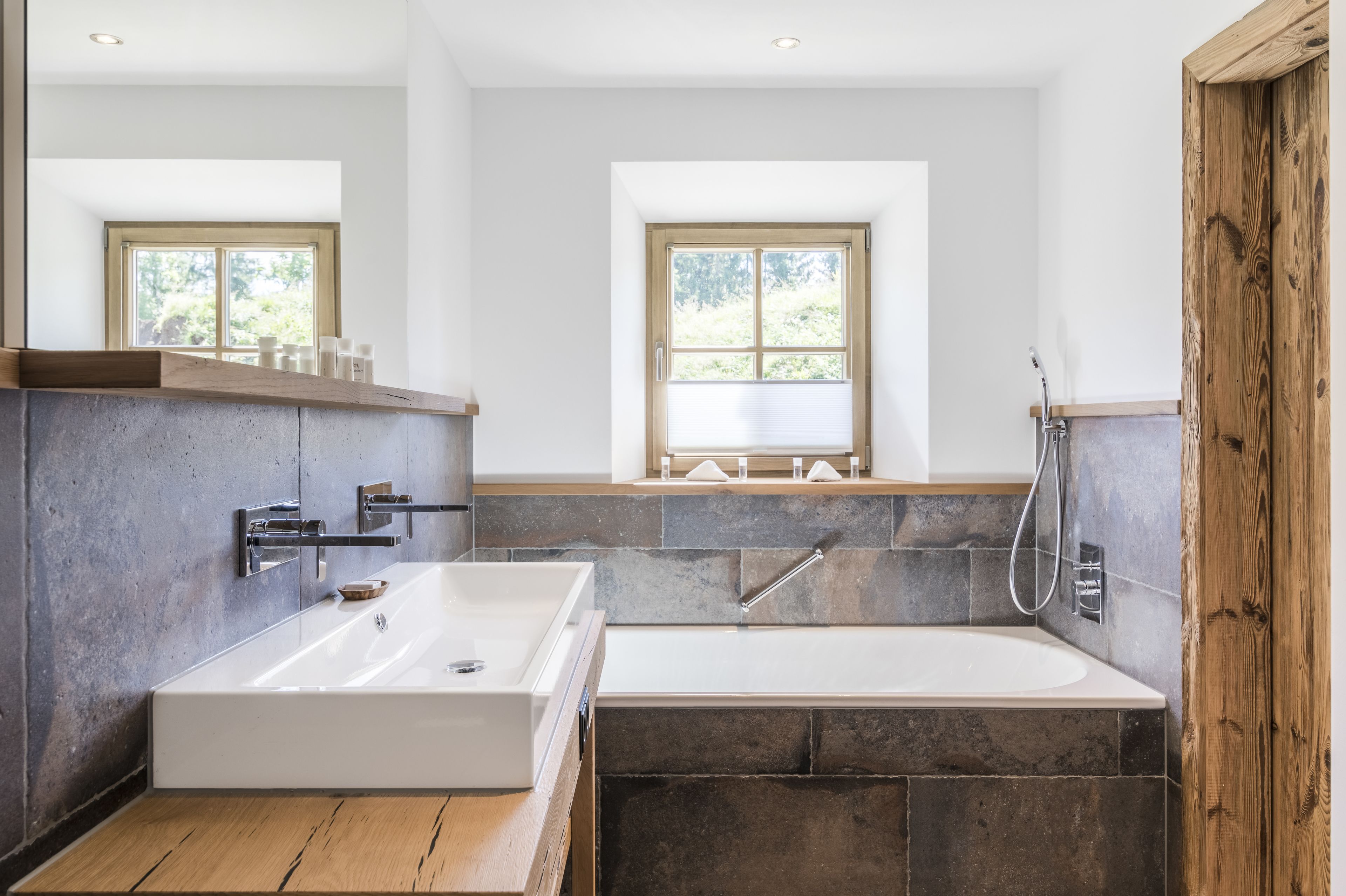 A modern bathroom with a large bathtub, a separate shower area, and a vanity with a sink. There are two windows providing natural light.