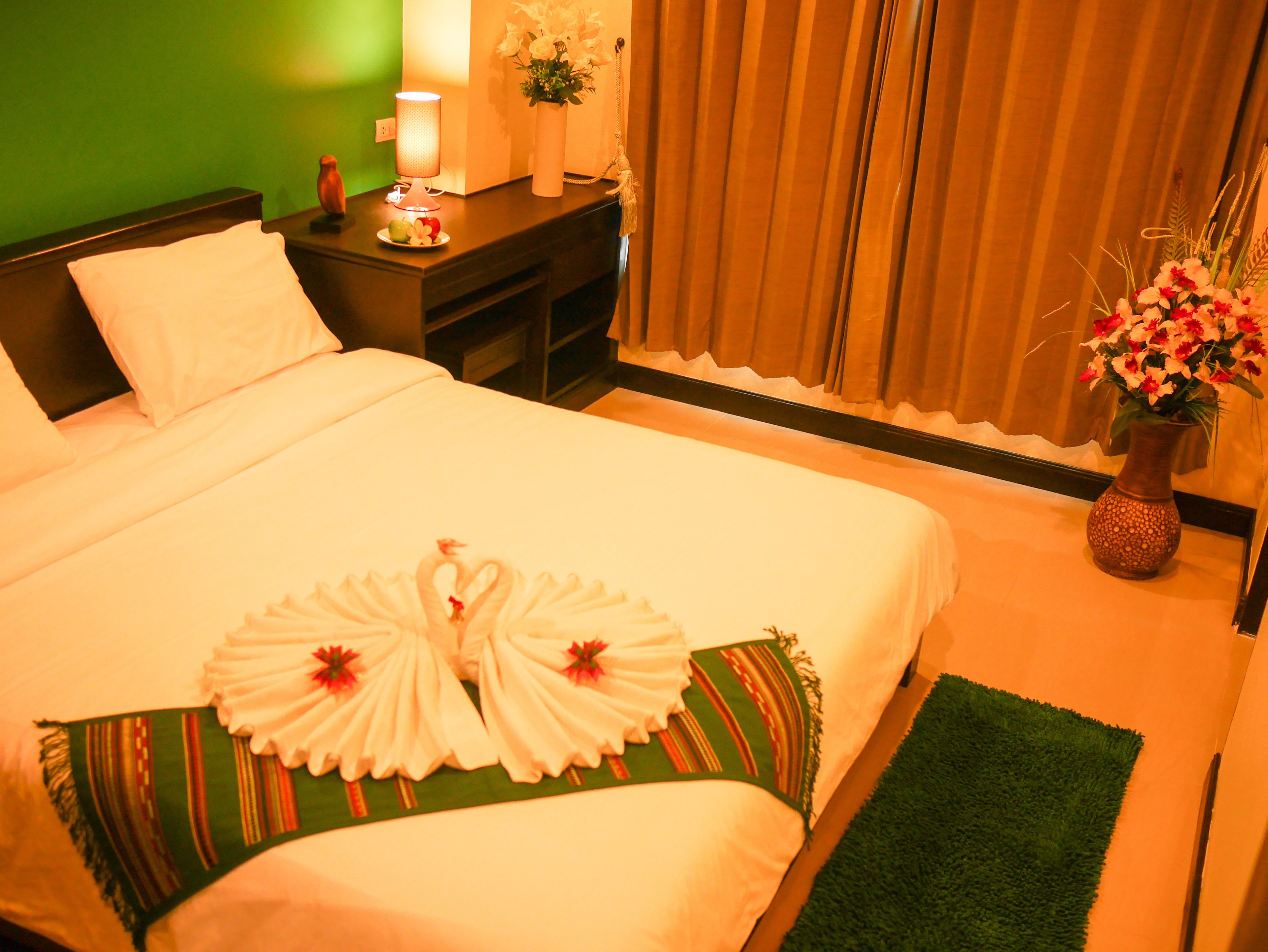 A neatly made bed with a heart-shaped towel arrangement, a bedside table with a lamp and a vase of flowers, and a green wall in the background.