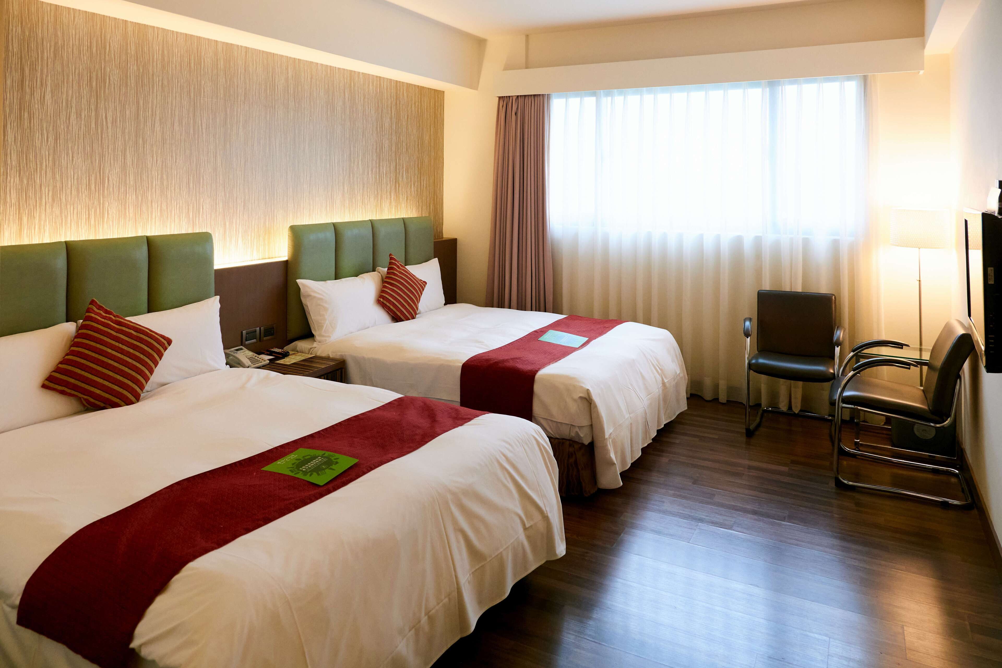 Habitación de hotel con dos camas, un escritorio, una silla y un ventanal con cortinas.