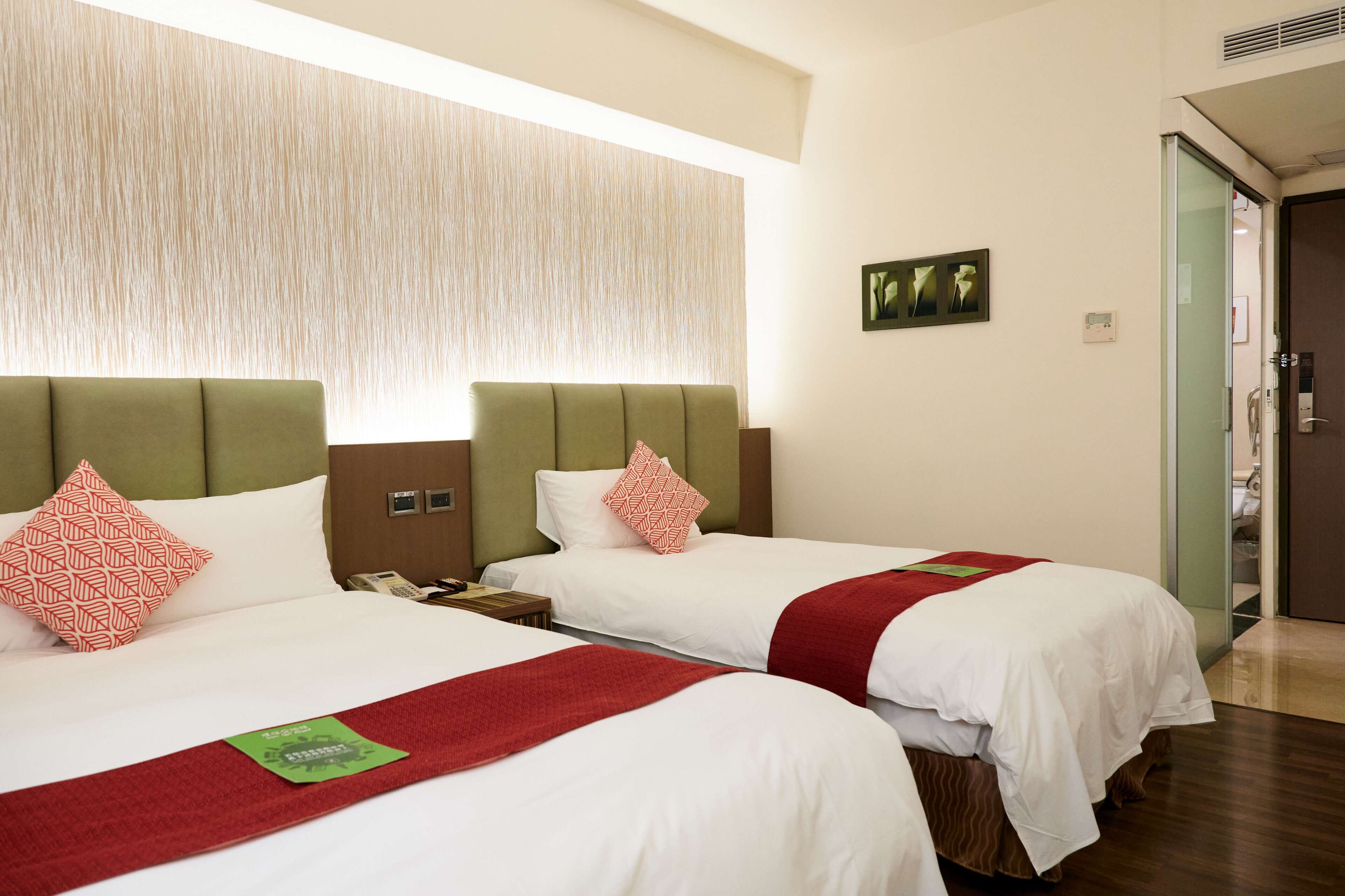 Habitación de hotel con dos camas, cada una con ropa de cama blanca y detalles en rojo, un cabecero verde y un ventanal grande con persiana texturizada.