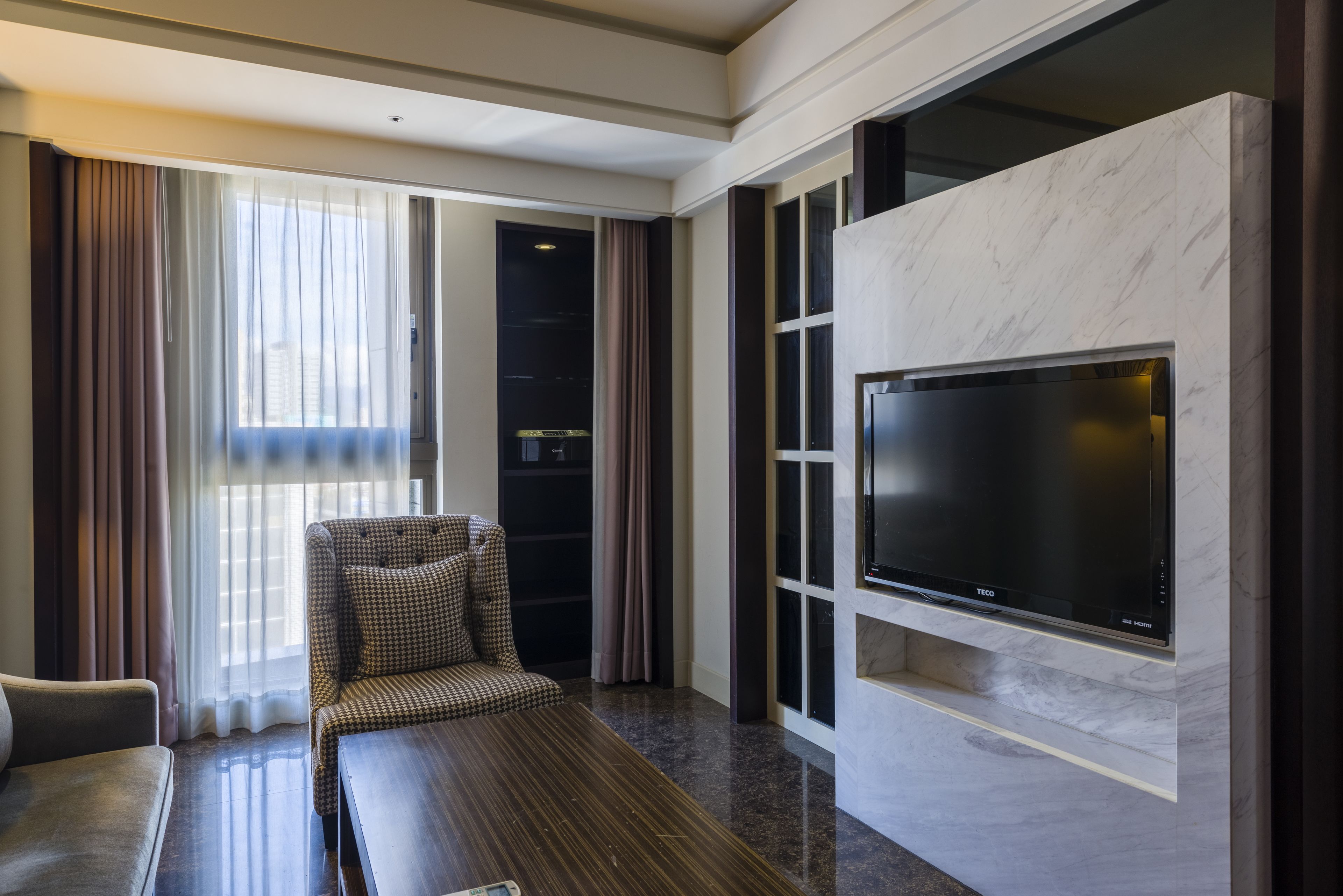A modern hotel room with a large flat-screen TV mounted on a marble wall, a comfortable sofa, a small table, and a view of the city through large windows with sheer curtains.