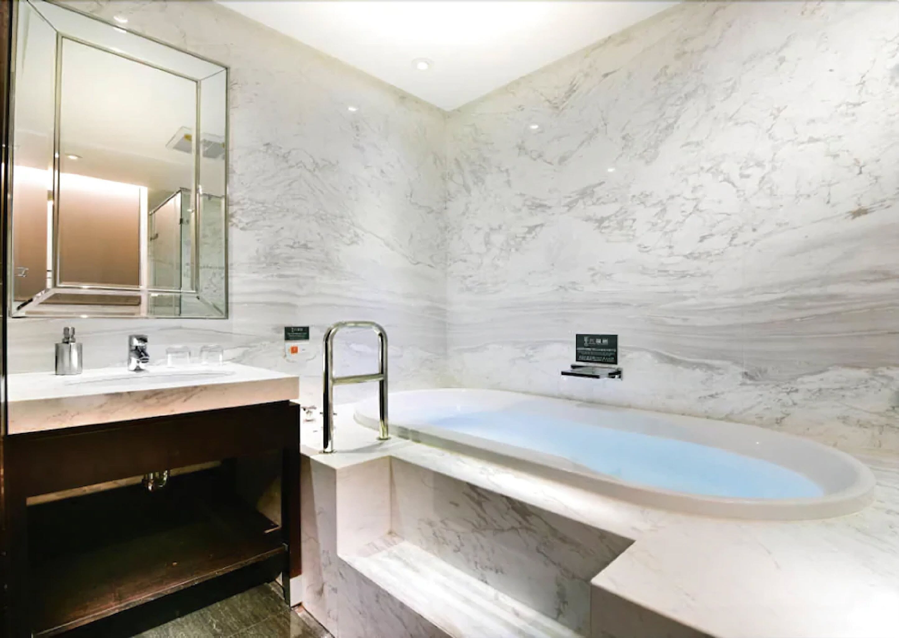 A modern bathroom with a marble wall, a freestanding bathtub, a vanity with a mirror, and a handrail.