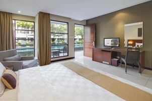 Una camera d'albergo con un letto, una scrivania, una sedia, una TV e ampie finestre con tende.