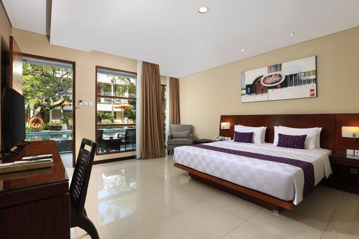 A hotel room with a large bed, a desk, a TV, and a view of an outdoor pool area.