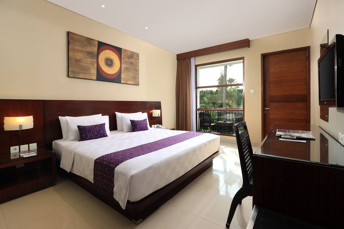 A hotel room with a large bed, a desk with a chair, a TV, and a window with a view of greenery.