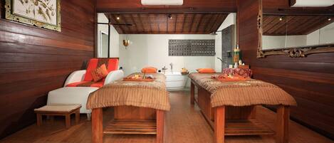 Sala de spa com paredes e teto de madeira, duas mesas de massagem, um sofá pequeno e um espelho fixado na parede.