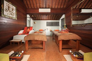 Sala de spa com paredes e teto de madeira, duas mesas de massagem, um sofá pequeno e um espelho fixado na parede.