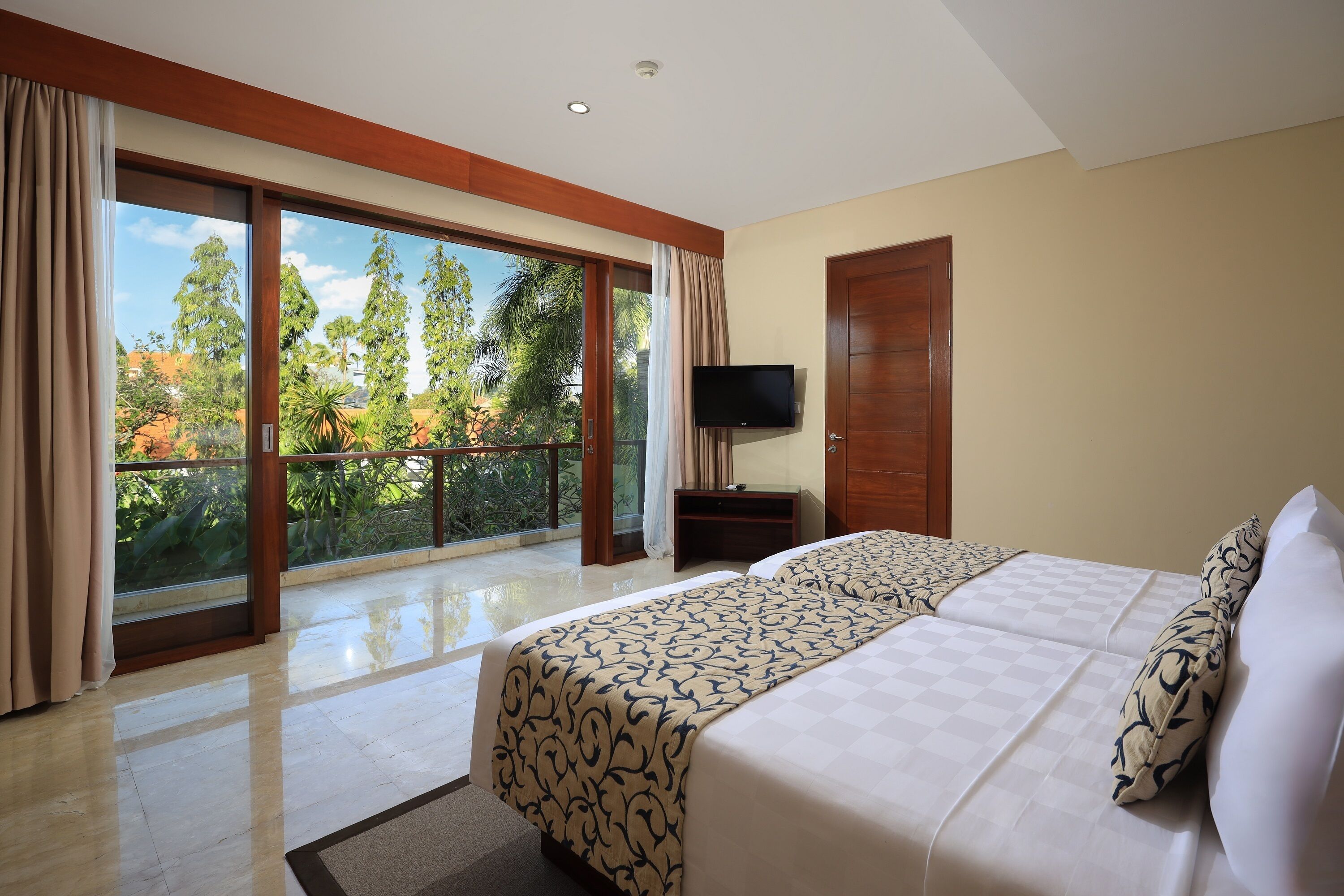 Habitación de hotel con dos camas, televisión y balcón con vistas a árboles y un cielo despejado.