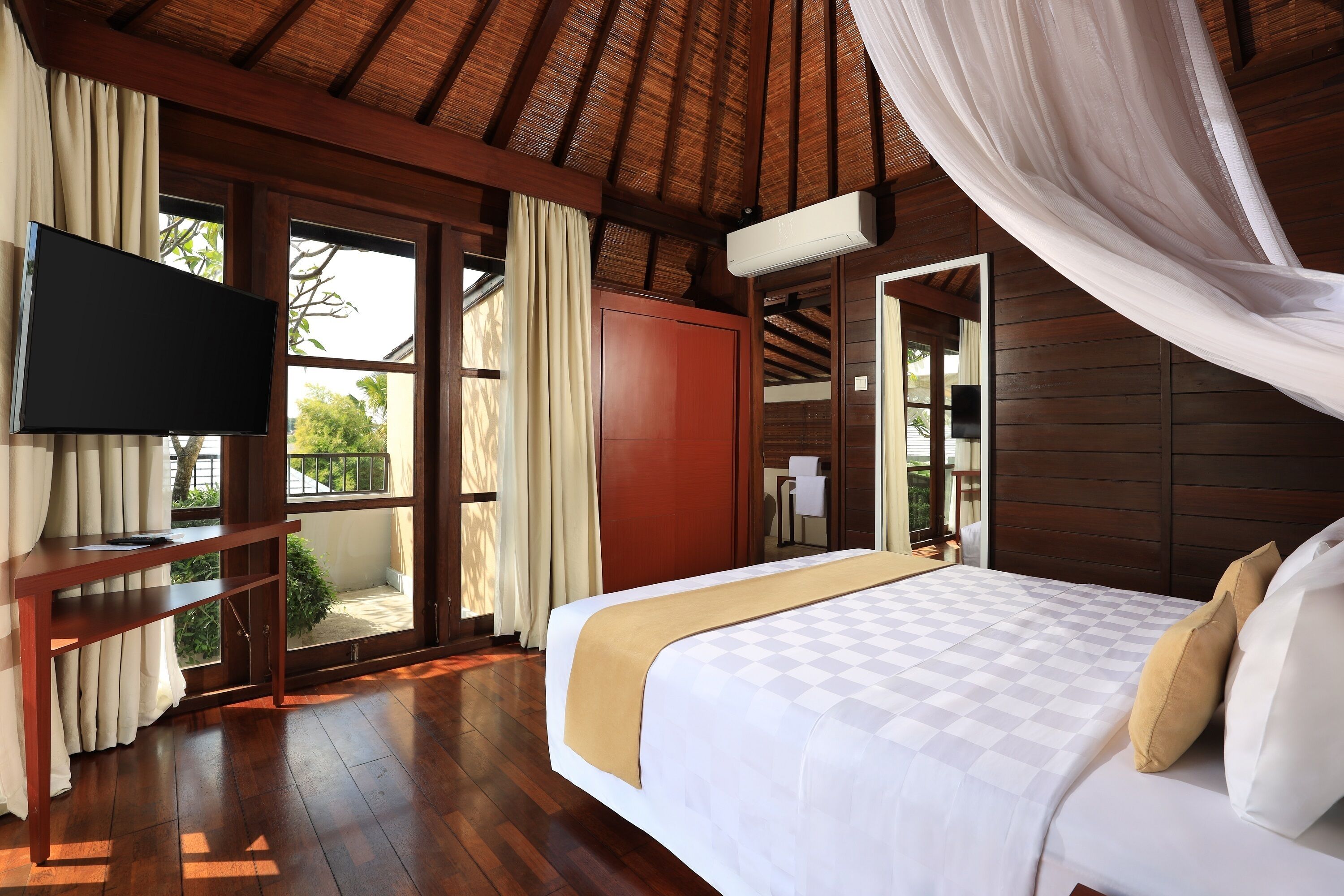 A bedroom with a large bed, a TV on a wooden console, and a view of the outdoors through a glass door.