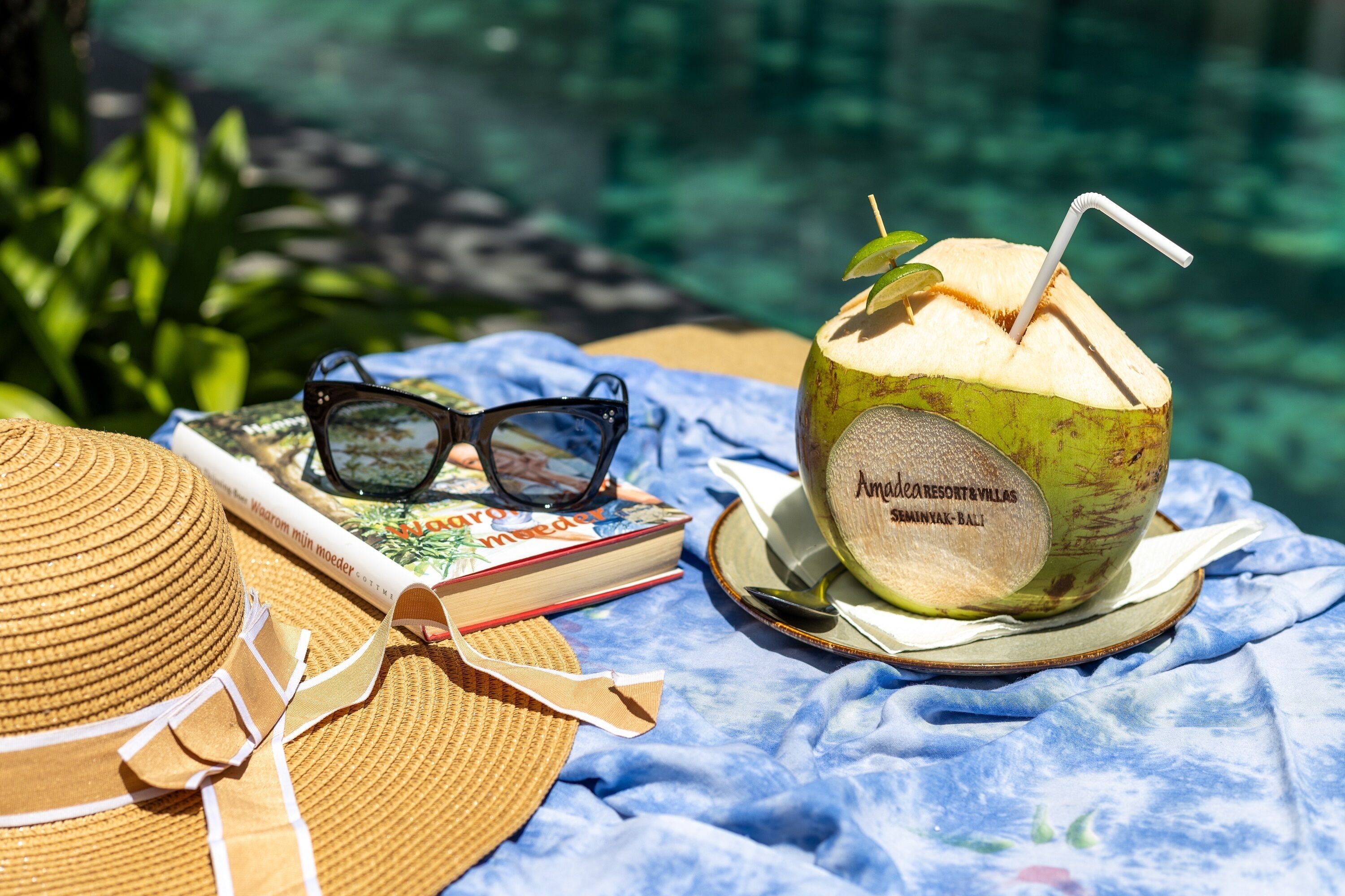 Une paille dans une noix de coco, des lunettes de soleil, un livre et un chapeau de paille posés sur un tissu bleu.