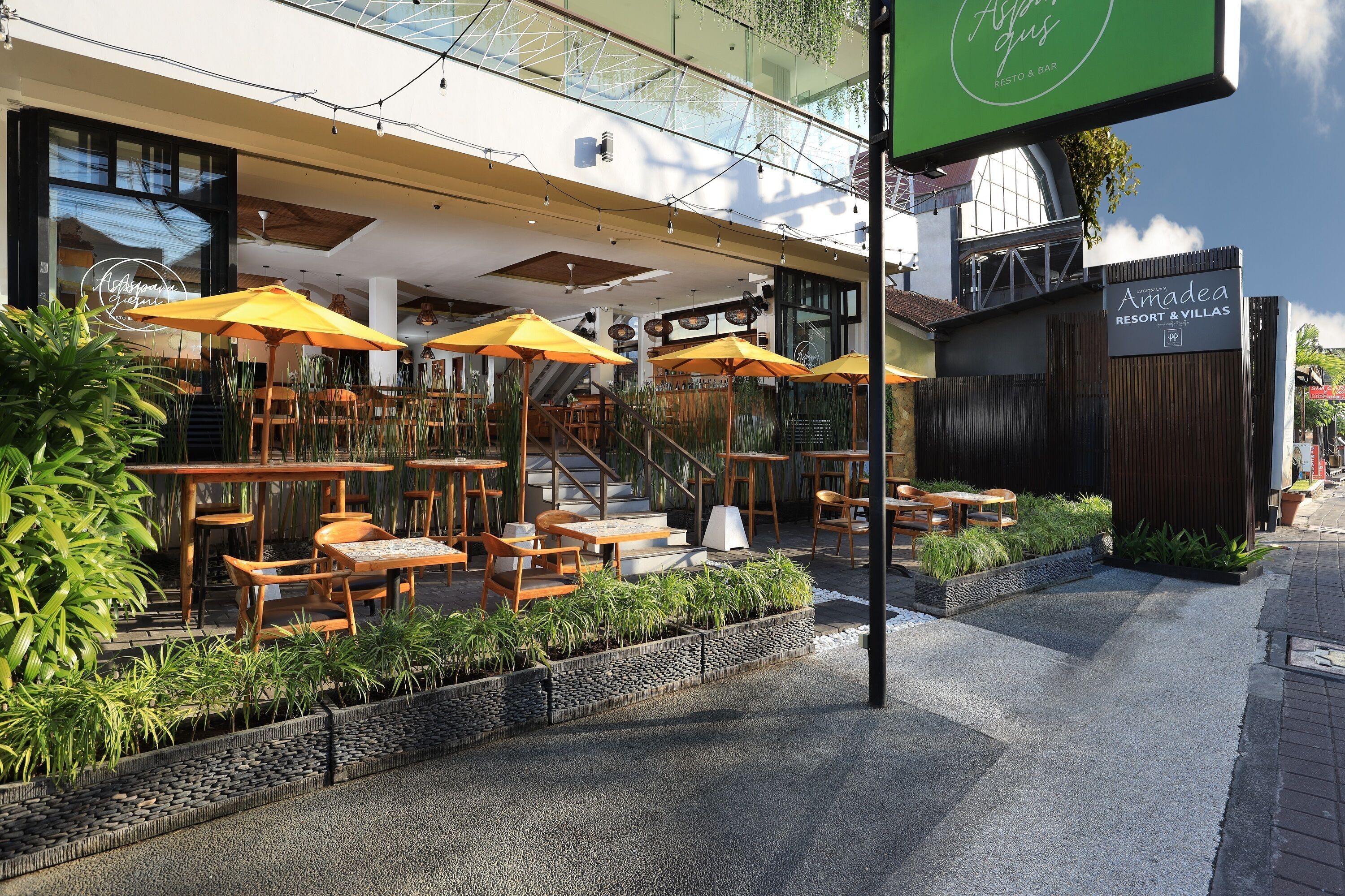 An outdoor seating area with wooden tables and yellow umbrellas, surrounded by planters and a modern building facade.